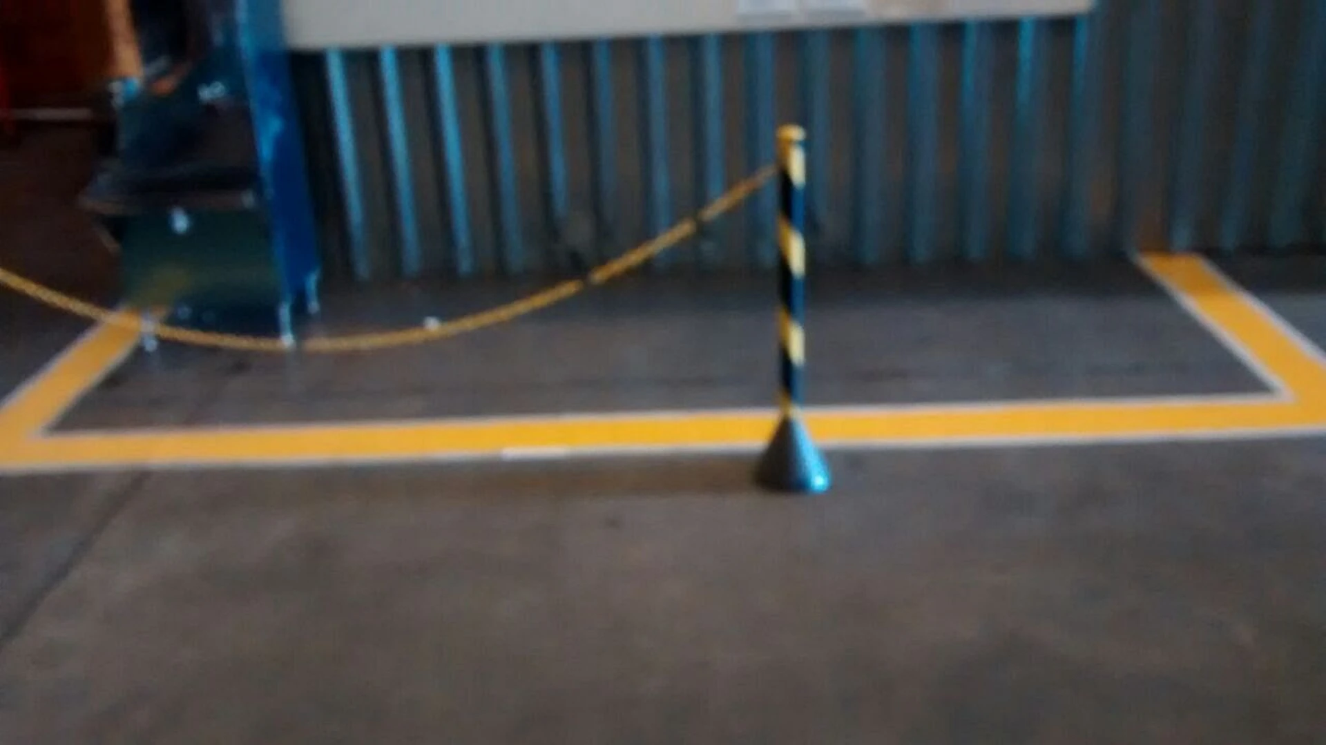 Área retangular delimitada por faixa amarela com cone de sinalização e parede metálica ao fundo