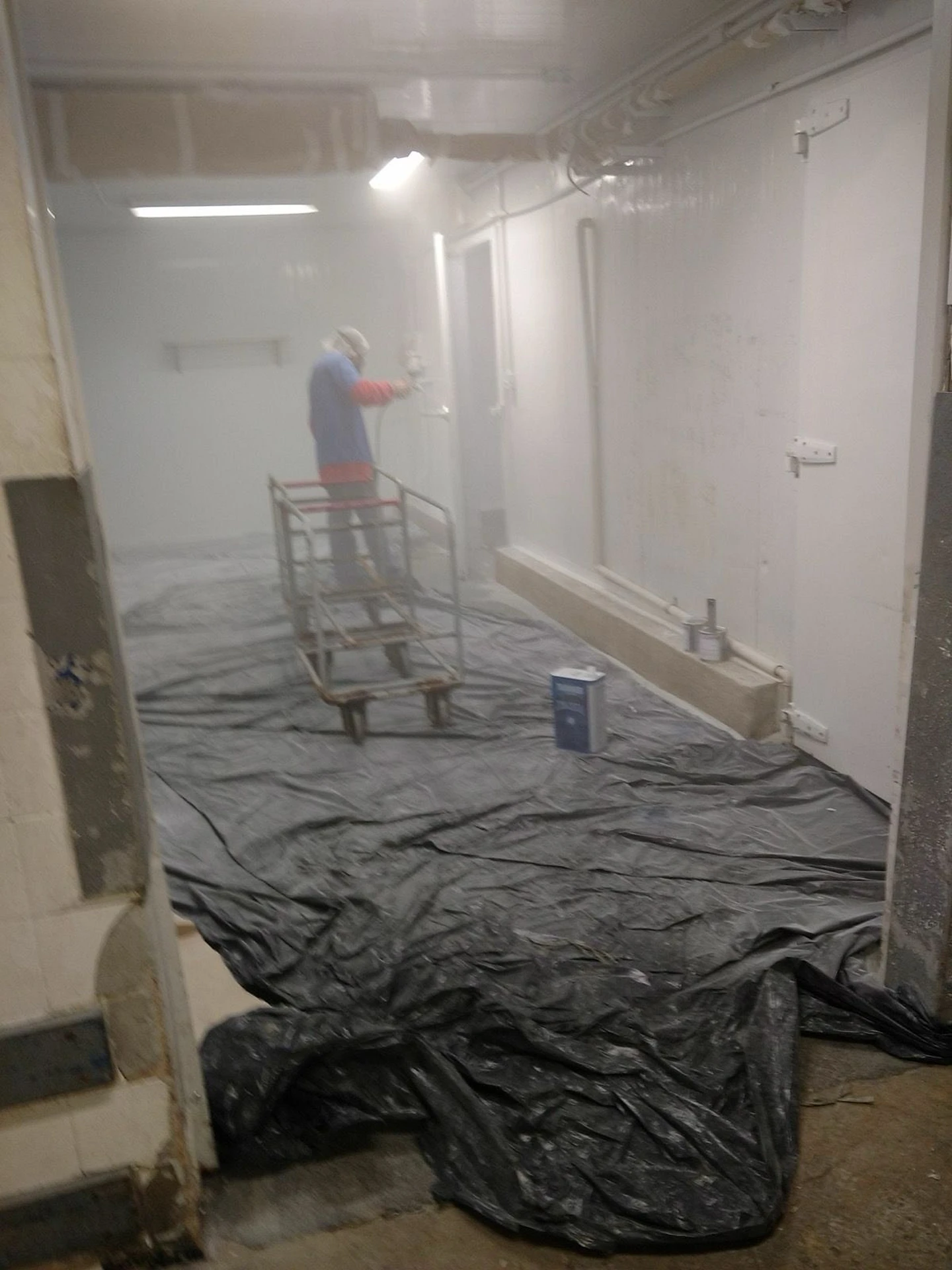 Trabalhador aplicando pintura com pistola no interior da câmara frigorífica com piso protegido por lona